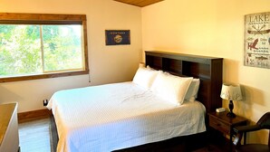 A bedroom with a wooden ceiling, a large window, a bed with white bedding, a nightstand with a lamp, and a wall sign that says "MONTANA". - Cozy Cabin With Mountain Views (Columbia Falls)