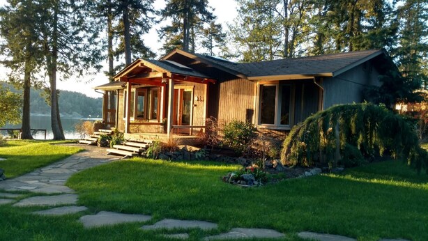 A wooden cabin with a stone pathway leading to the entrance, surrounded by lush greenery and tall trees.