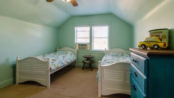 Two single beds with matching headboards, a small table, and a dresser are featured in this room, along with a ceiling fan and windows with blinds.