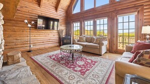 A log cabin interior with a wooden ceiling, large windows, a sofa, a table, and a fireplace. - Stunning Mountain Views on 8 Acres with Hot Tub, sleeps 10 (Saluda)