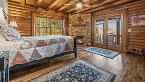 A log cabin bedroom with a bed, a stone fireplace, wooden floors, and a balcony with glass doors. - Stunning Mountain Views on 8 Acres with Hot Tub, sleeps 10 (Saluda)