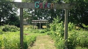 Ein hölzerner Bogen mit der Aufschrift 'Garden', der zu einem üppigen Garten mit verschiedenen Pflanzen und einer kleinen Struktur im Hintergrund führt.