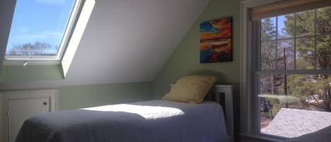 Um quarto com claraboia, uma cama com edredom branco, uma janela com vista para árvores e um quadro na parede.
