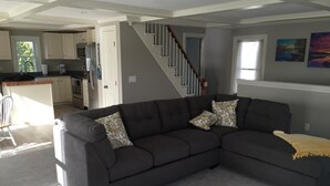 Ein modernes Wohnzimmer mit einem dunkelgrauen Ecksofa, einer gelben Decke und gemusterten Kissen. Eine Treppe führt zu einer offenen Küche mit weißen Schränken und einem Essbereich.