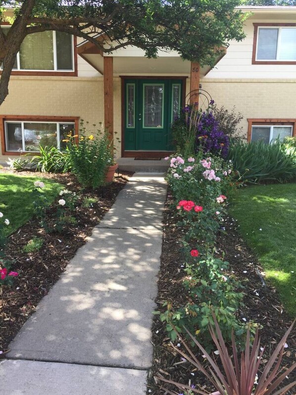 Un bâtiment à deux étages avec une porte d’entrée verte, un porche abrité et un jardin bien entretenu où poussent une grande variété de fleurs et de plantes.