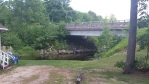 A concrete bridge over a calm river, surrounded by greenery and a gravel path.