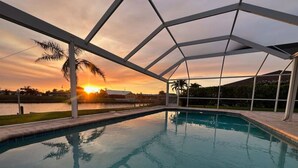 Une piscine dotée d’un toit transparent, entourée d’une pelouse et de palmiers, avec un coucher de soleil en arrière-plan.