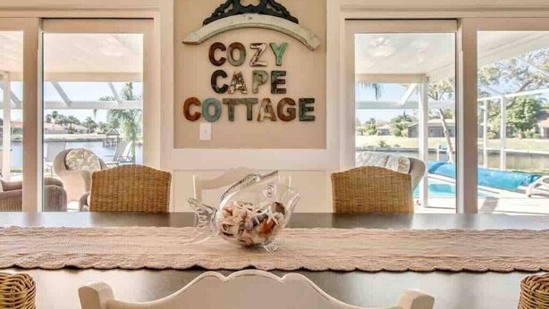 Un coin repas avec des chaises en osier, une table avec un set de table et une pancarte indiquant : « COZY CAPE COTTAGE ».