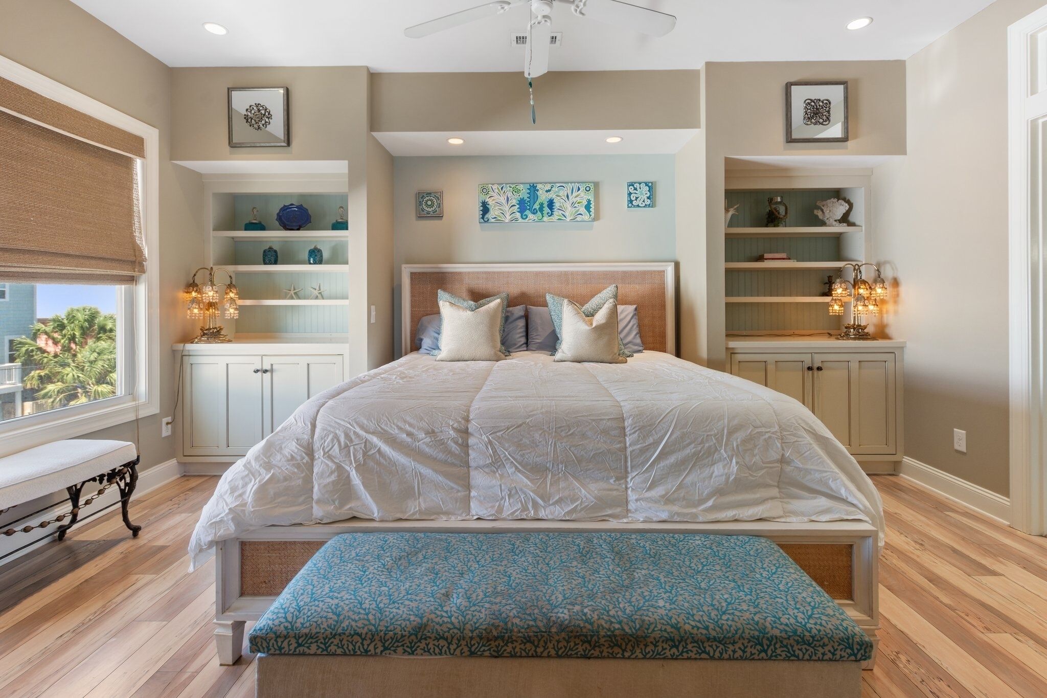 Un dormitorio con una cama grande, estanterías empotradas con objetos decorativos, un ventilador de techo y una ventana con vistas a la vegetación.