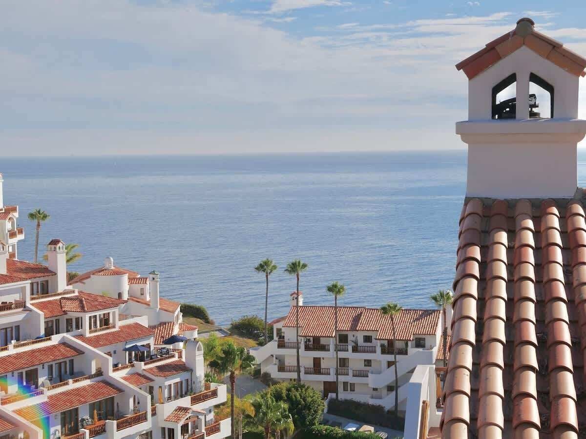 A coastal hotel with multiple balconies, a view of the sea, and palm trees.