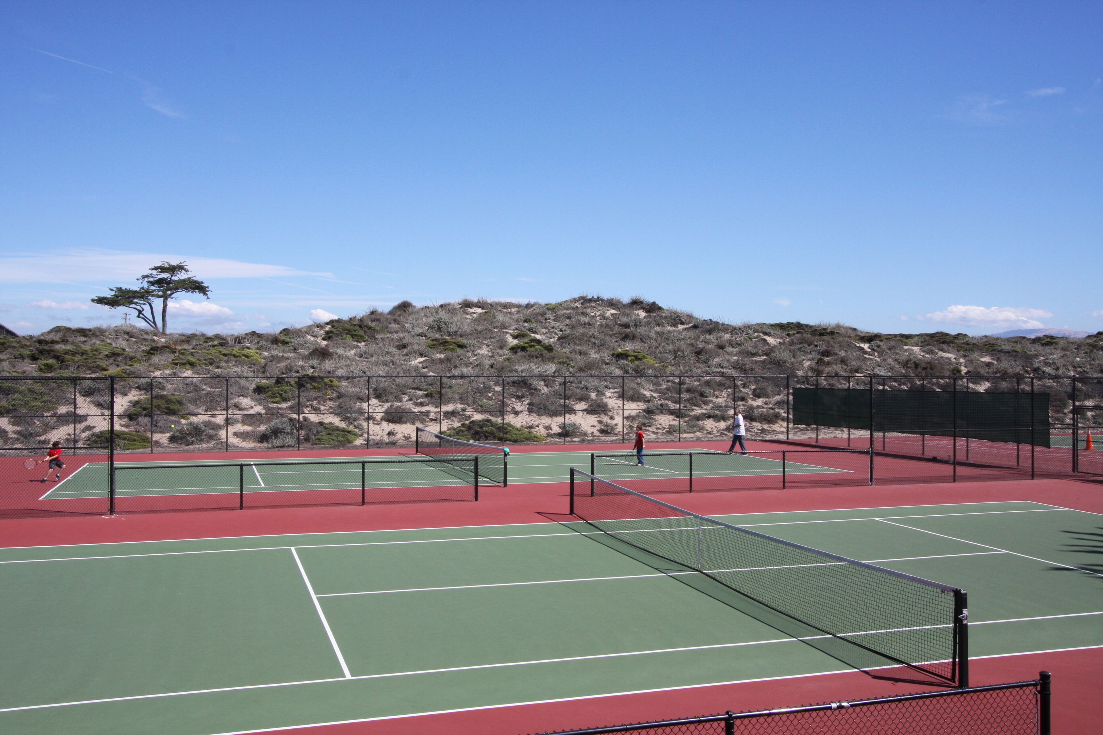 Pista de tenis exterior con varias canchas, rodeada por una valla y un paisaje montañoso.