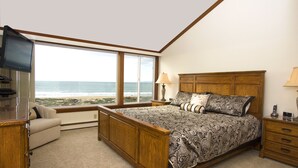 A bedroom with a large window offering a view of the beach, a wooden bed, a bedside table, and a TV mounted on the wall.