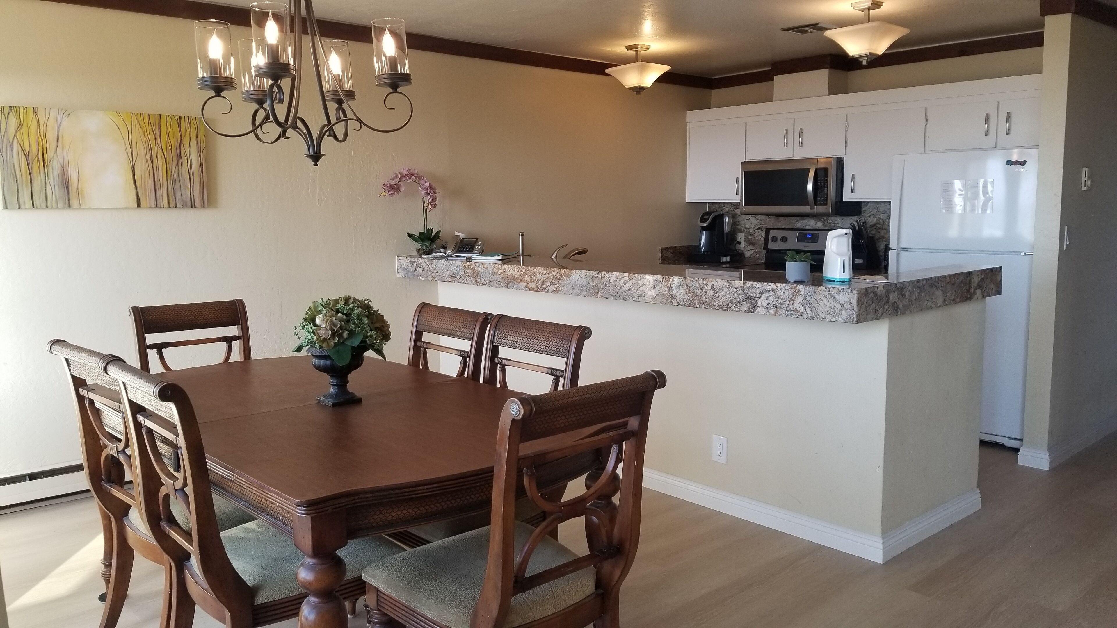 A dining area with a wooden table and chairs, a chandelier, and a kitchen with white cabinets and appliances.