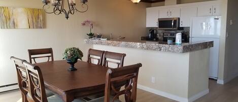 A dining area with a wooden table and chairs, a chandelier, and a kitchen with white cabinets and appliances.
