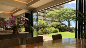 Un hall d’accueil d’hôtel avec un comptoir de réception en bois, des chaises en osier et une grande fenêtre donnant sur un jardin luxuriant et un bâtiment situé de l’autre côté de la rue.