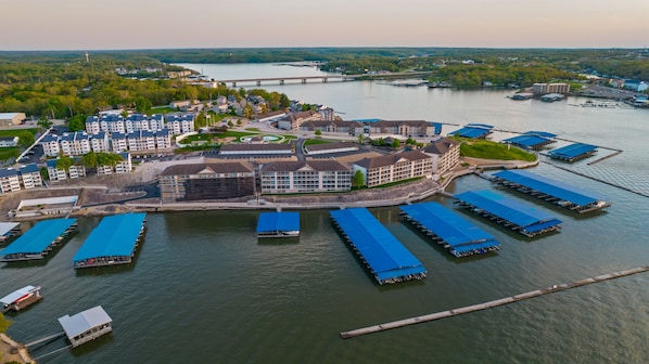An aerial view of a waterfront hotel with multiple docked boats, a bridge in the background, and a forested landscape.