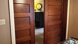 Una porta in legno che dà su una stanza con una ventola a soffitto, un tavolino con una rivista e una pianta su uno scaffale.