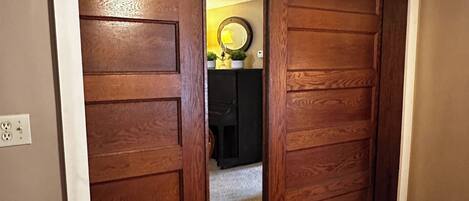 A wooden door leading to a room with a ceiling fan, a small table with a magazine, and a plant on a shelf.