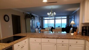 A kitchen with white cabinets, a granite countertop, and a built-in stove. There is a dining area with a view of the ocean. - Amazing Beachfront Destin Condo With Ocean Views from All Rooms (Destin)