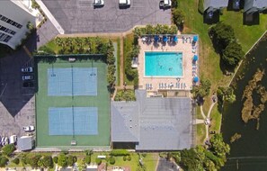 Vue aérienne d’une propriété comprenant un court de tennis, une piscine avec des chaises longues et un espace de stationnement.