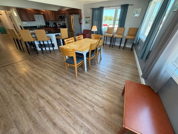 A modern kitchen with wooden cabinets, a dining area with a wooden table and chairs, and a view of the outside through large windows.