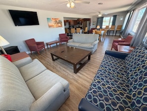 A living room with a sofa, armchairs, a coffee table, and a TV. There is a kitchen area in the background.