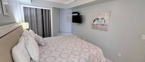 Un dormitorio con cama, ventilador de techo, ventana con cortinas, televisor montado y una obra de arte en la pared.