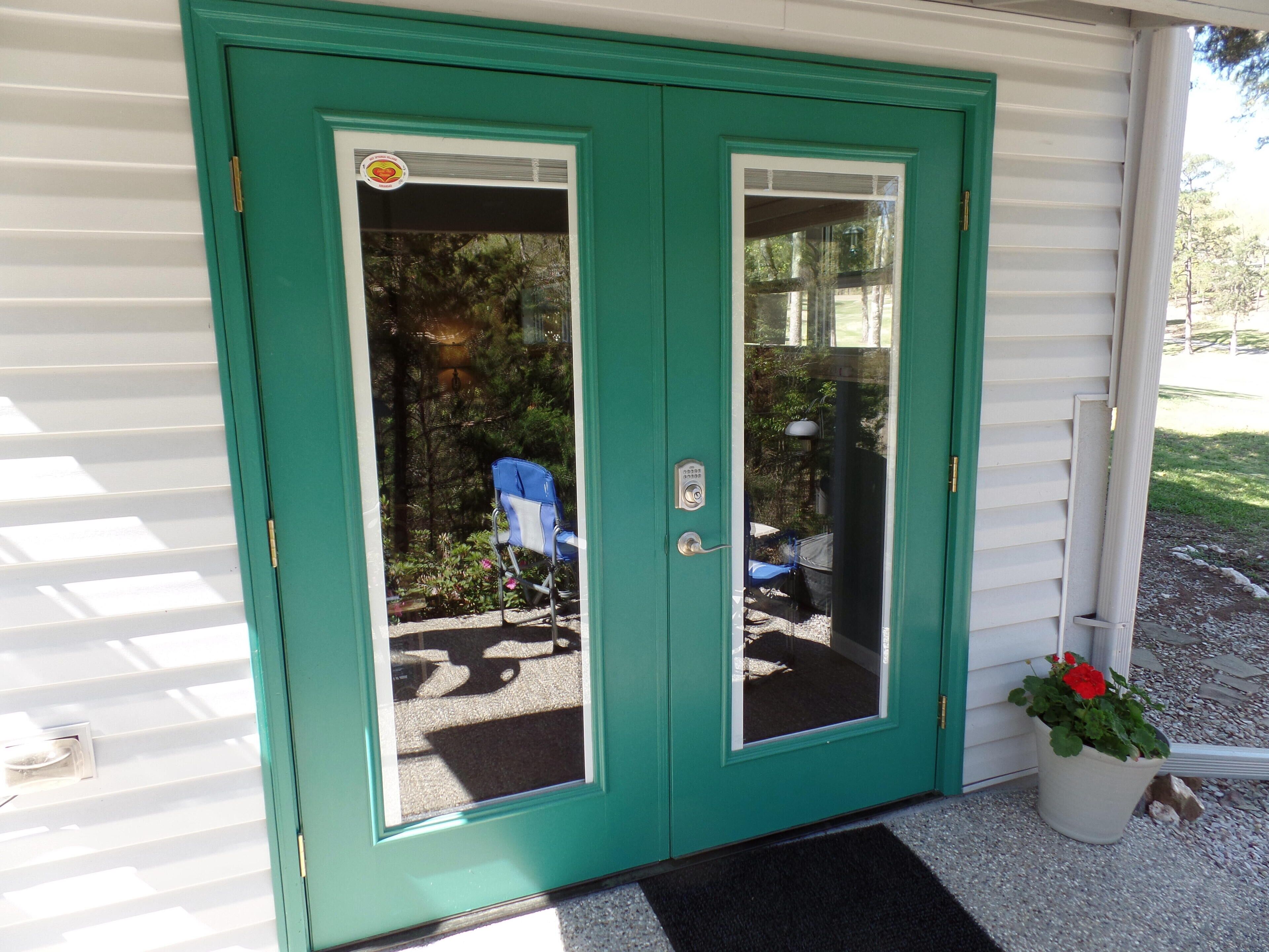 Una puerta doble color turquesa con un panel de vidrio, una silla azul y una planta en maceta afuera.