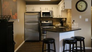 A kitchen with white cabinets, a stainless steel refrigerator, a microwave, an oven, and a clock on the wall. There are three stools at the island. - Don't Click Here UNLESS You Want The Best Value In Southern Utah! (St George)