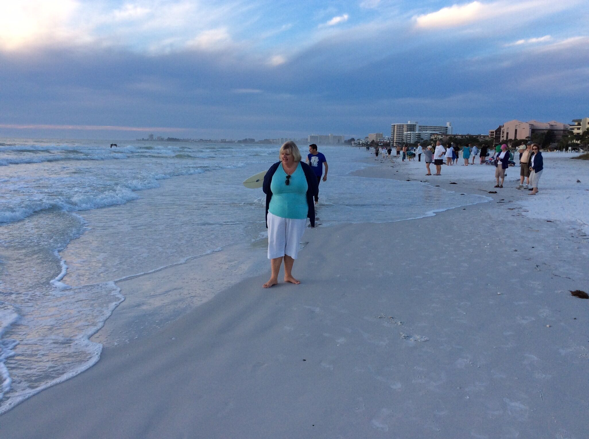 A beach with people walking and standing, waves crashing onto the shore, and buildings in the background.