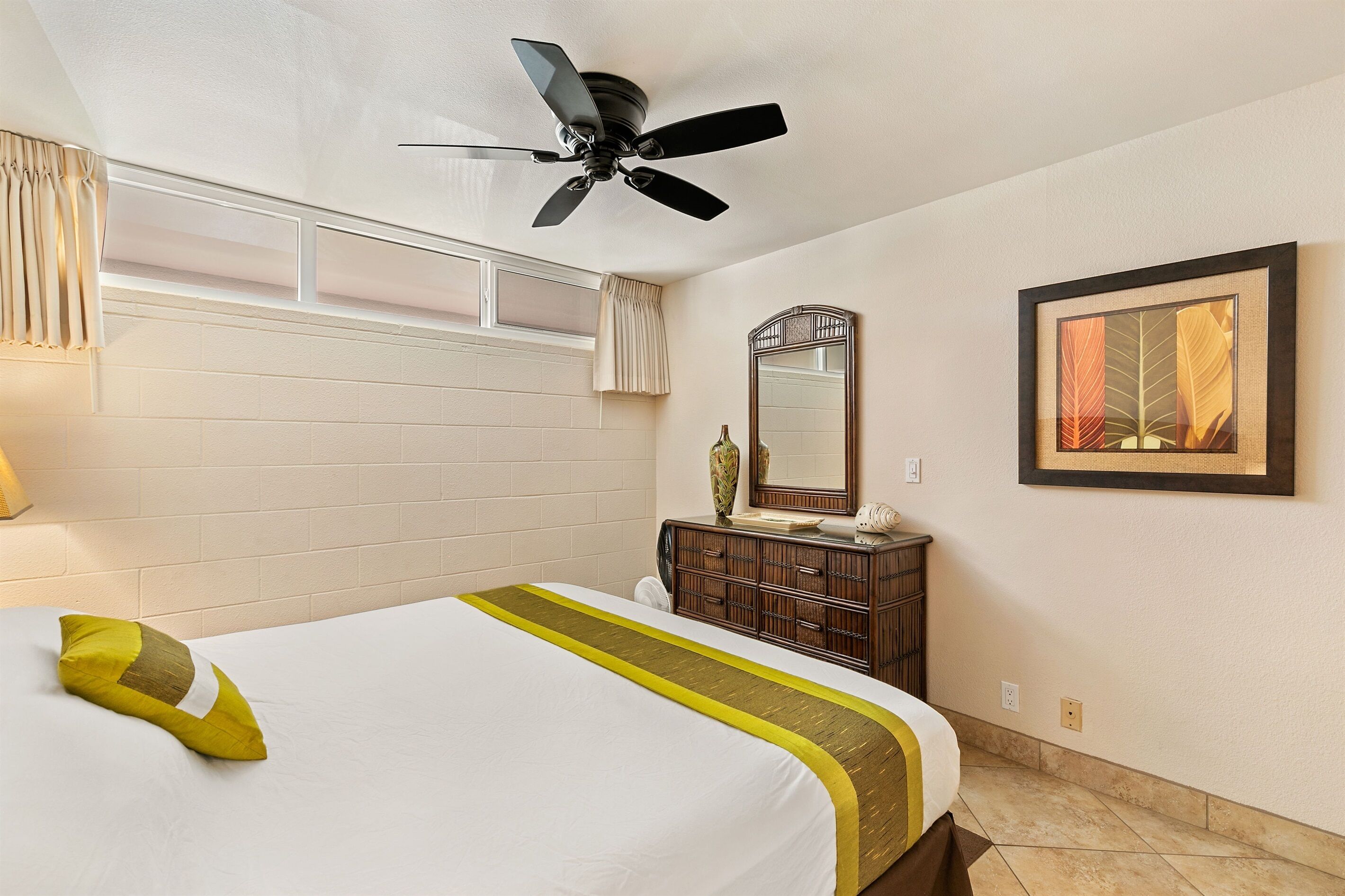A hotel room with a bed, a ceiling fan, a dresser, a mirror, and a framed picture on the wall.