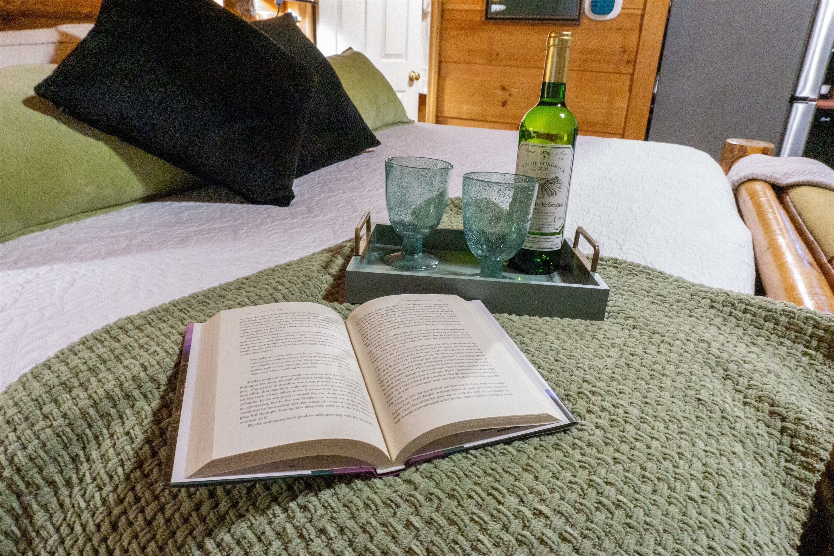 Un letto con una coperta verde, un libro, due bicchieri e una bottiglia su un vassoio.