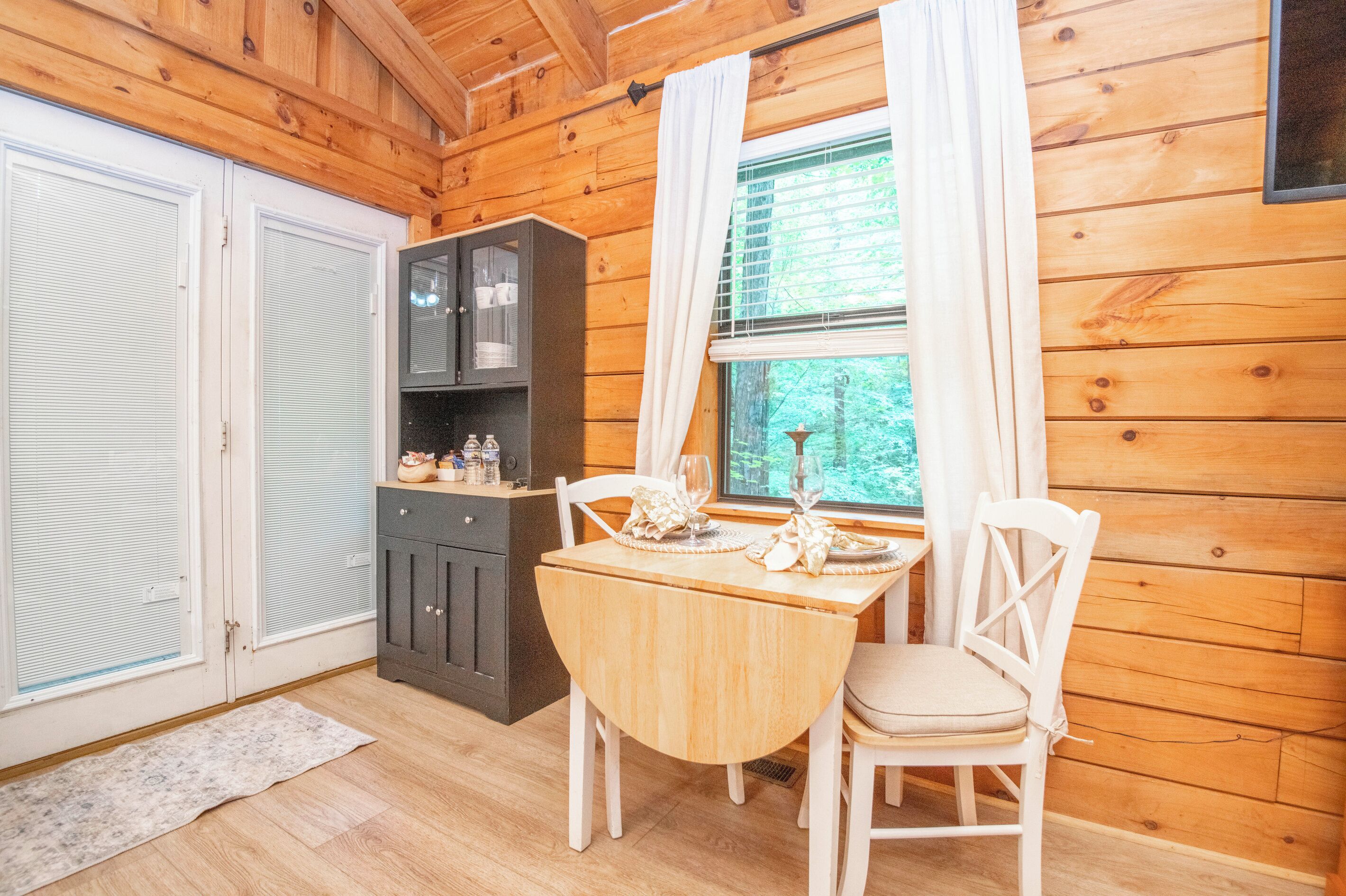 A wooden interior with a dining table set for two, a cabinet with dishes, and a window with curtains.