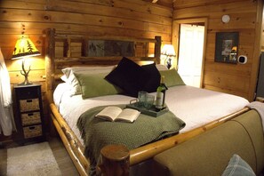 A wooden cabin bedroom with a bed, pillows, a bedside table with a lamp, a small table with a bottle and glasses, and a bookshelf. - Private Log Cabin with Hot Tub (Gatlinburg)