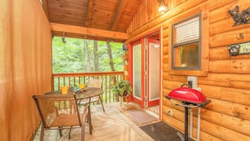 A wooden cabin with a balcony, a dining table with chairs, a red grill, and a window with a bear decoration.