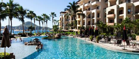 A resort pool with clear blue water, surrounded by palm trees and a multi-story building with balconies.