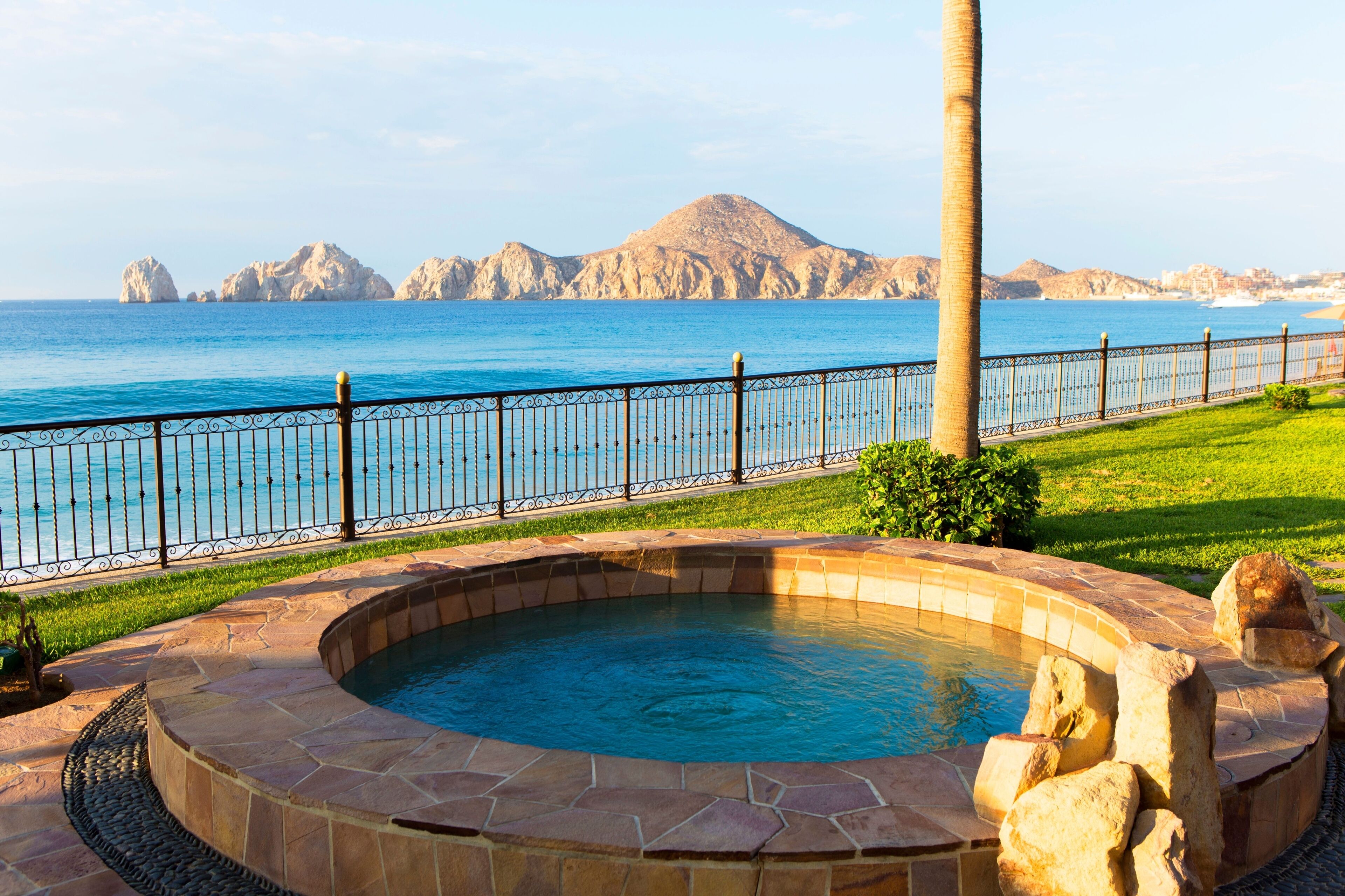 Una piscina con borde de piedra, cerca metálica y vista al mar y montañas de fondo.