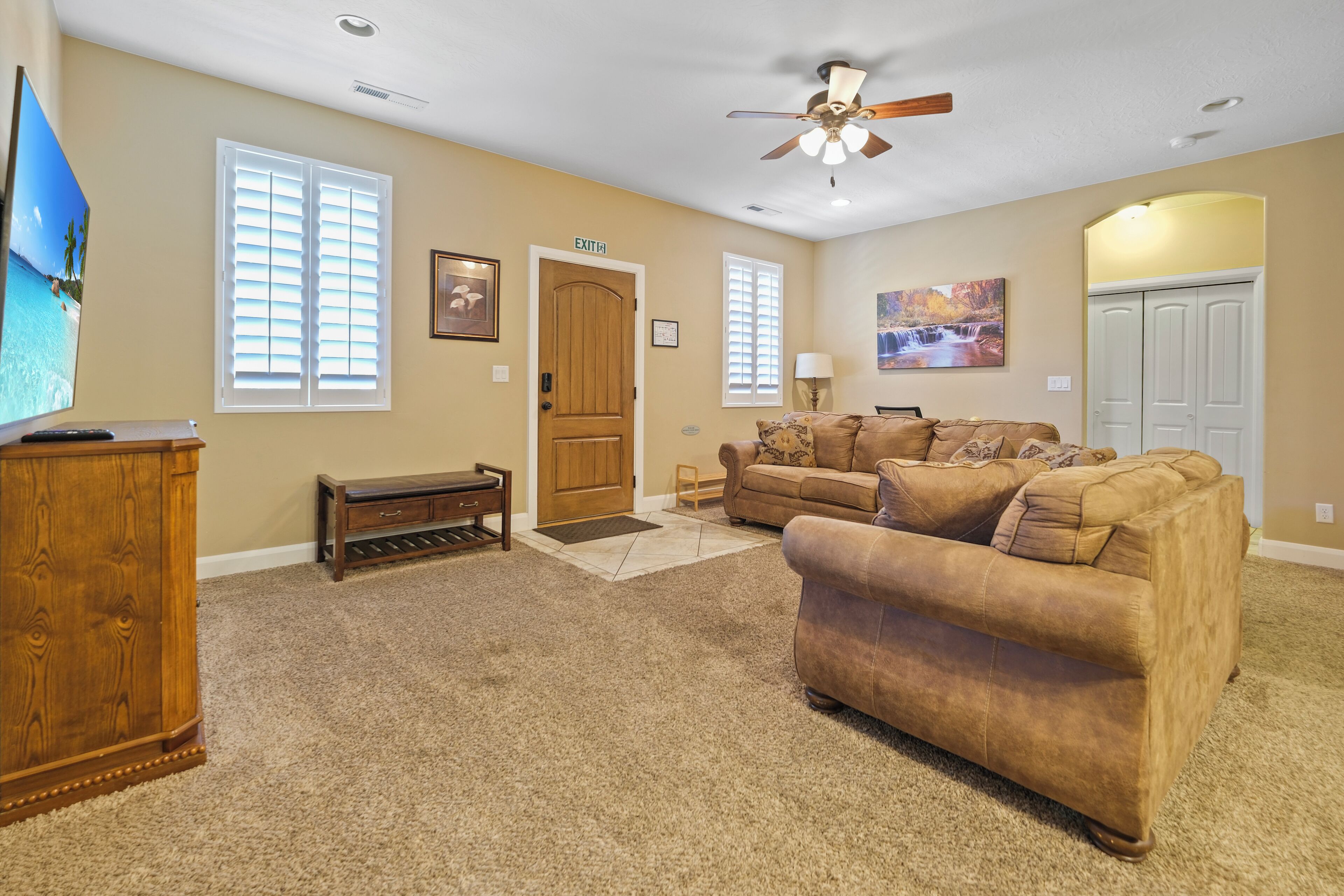 A living room with beige walls, a ceiling fan, a wooden door labeled "EXIT", a brown sofa, a wooden cabinet, and a television.