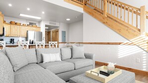 Una sala de estar moderna con un sofá seccional gris, una escalera de madera y una cocina al fondo.