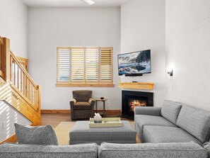 Una sala de estar con chimenea, televisor montado encima, un sofá, una butaca y una ventana con persianas.