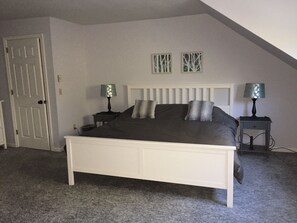 Un dormitorio con una cama blanca, dos mesitas de noche con lámparas, una puerta y un techo inclinado.