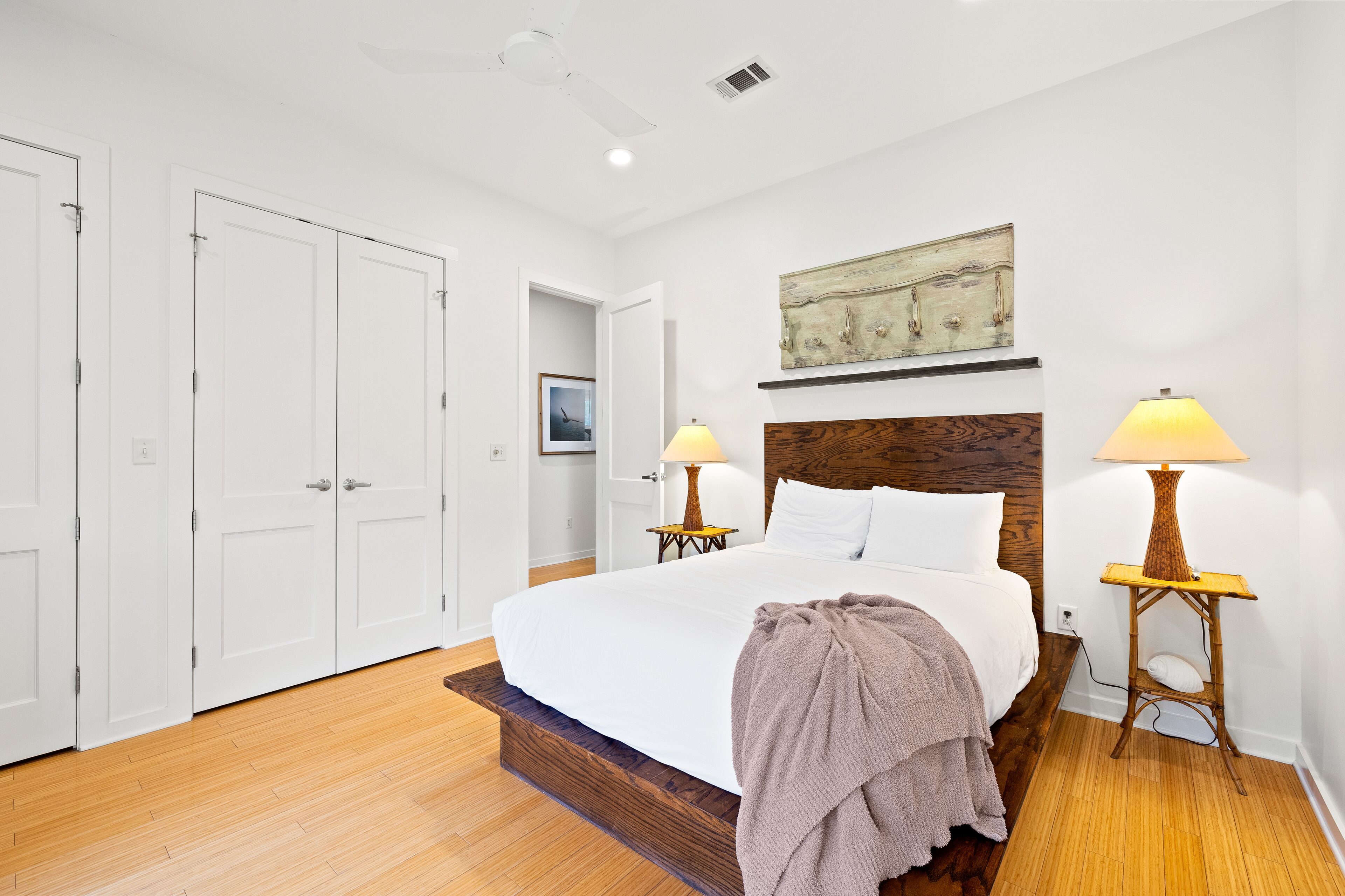 A bedroom with a wooden bed frame, white bedding, two bedside lamps, a wooden headboard, and a painting above the bed.