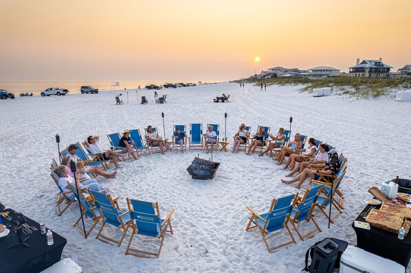 Guests gather around a central fire pit on the wide, sandy beach, surrounded by well-maintained facilities and ample parking.