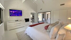 Un dormitorio amplio con una cama grande, un televisor instalado en la pared, un ventilador de techo y un ventanal corredero que da a un balcón.