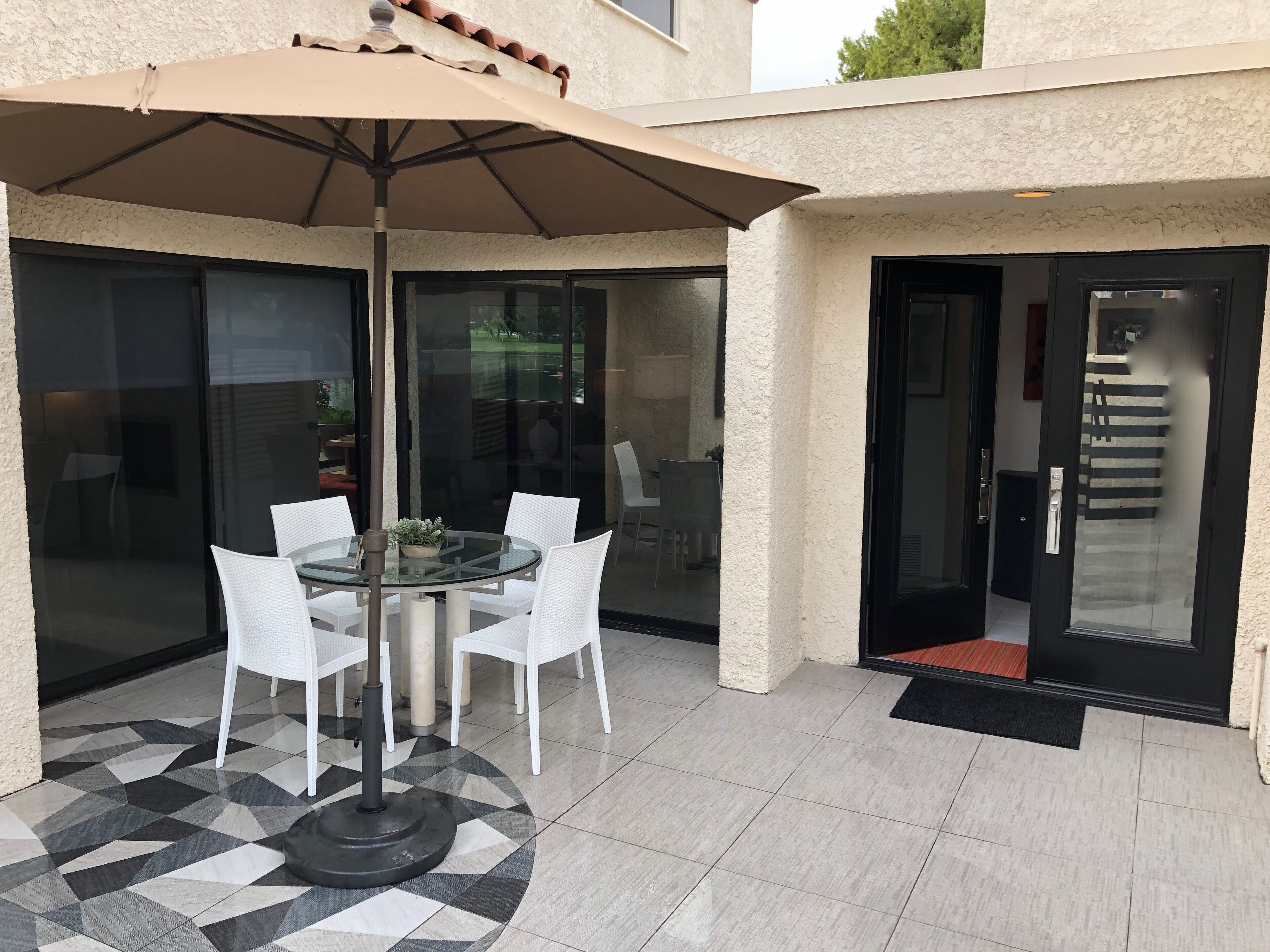 An outdoor patio with a round glass table and white chairs, a beige umbrella, and a patterned floor.