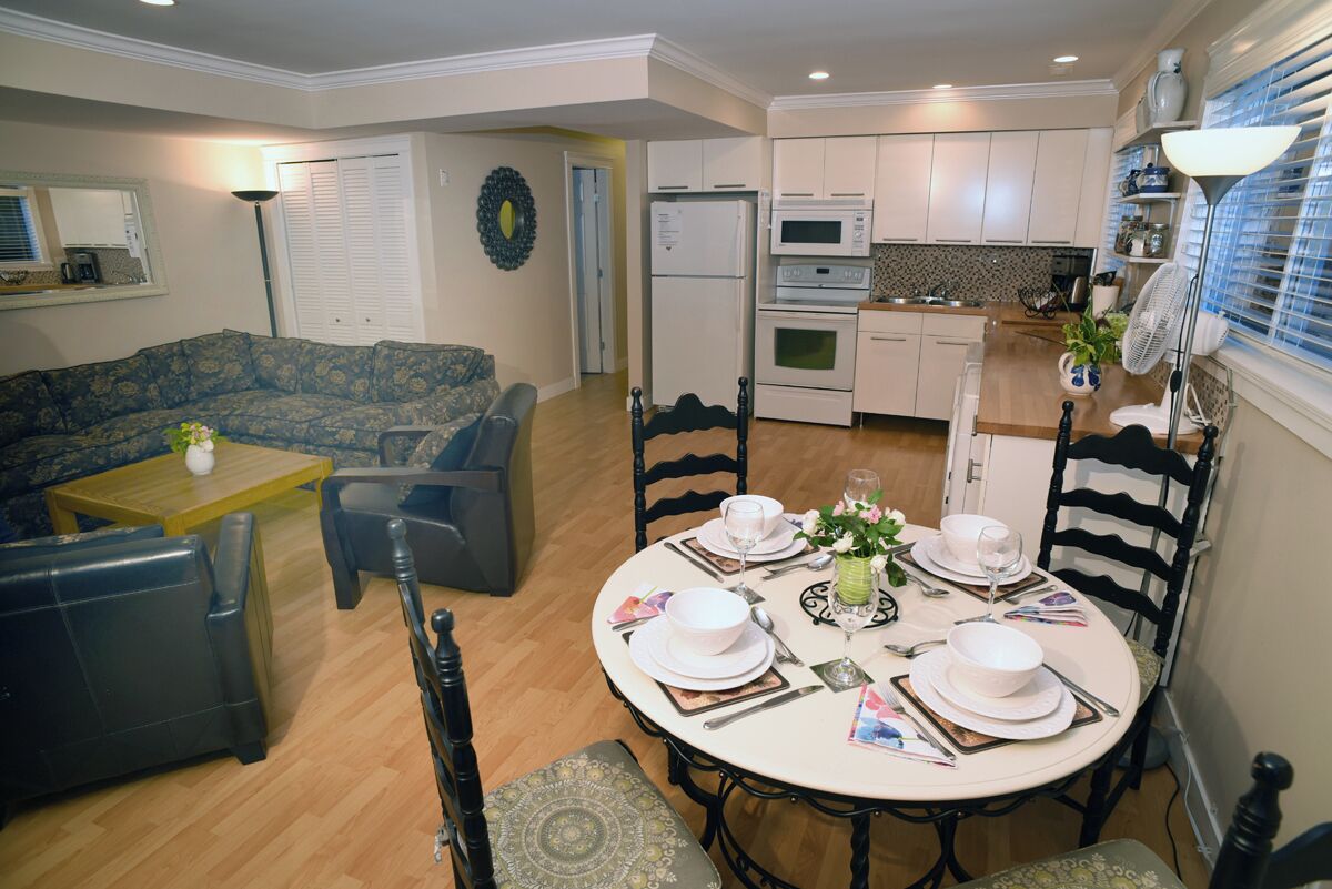A dining area with a round table set for four, surrounded by black chairs, in a room with a sofa and a kitchen in the background.