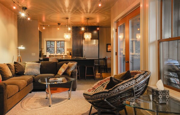 A cozy living room with a sofa, a glass coffee table, and a patterned armchair. A kitchen with a refrigerator and a window is visible in the background.