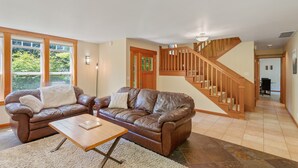 Un vaste salon agrémenté de deux canapés en cuir marron, d’une table basse en bois et d’une grande fenêtre donnant sur un paysage verdoyant. Un escalier en bois avec une rampe est visible au fond.