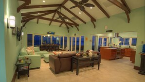 A spacious living room with a brown sofa, a glass coffee table, and a dining area with a table and chairs. The room has a high ceiling with exposed beams and a ceiling fan. - Location!!! no car needed, Luxury Oceanfront, Our home on HGTV (Nassau)