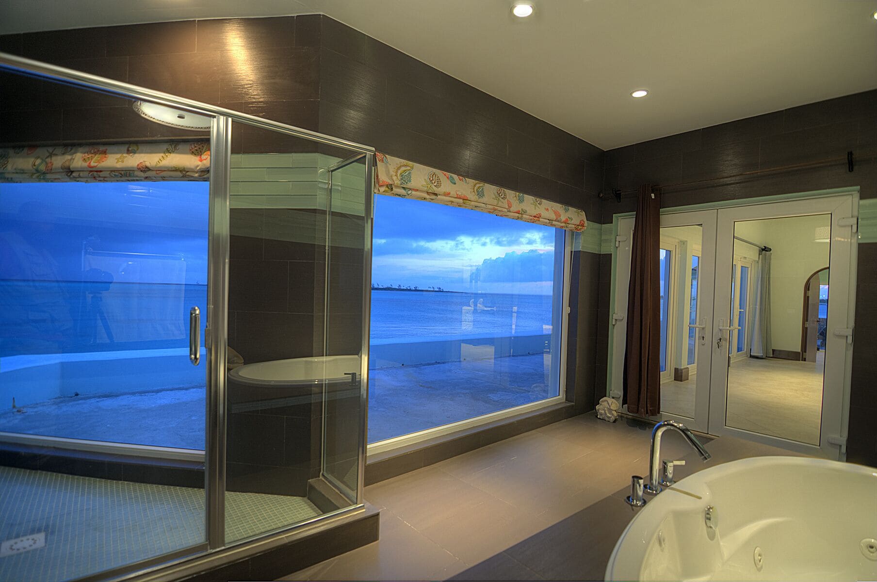 A modern bathroom with a glass-enclosed shower, a large bathtub, and a view of the sea.